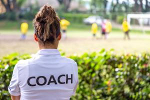Back view of a female soccer, football, coach in white coach shirt watching her team play at an outdoor football field